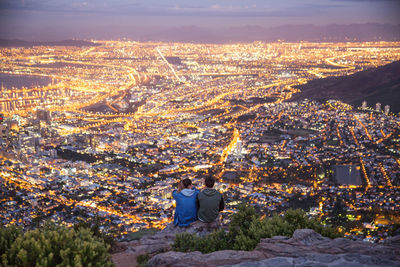 Rear view of man and woman sitting on mountains against illuminated city at dusk