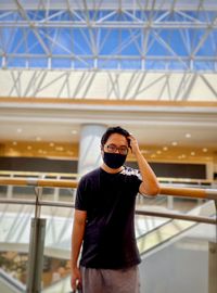 Low angle view of young man in eyeglasses and face mask standing against architecture and skylight.