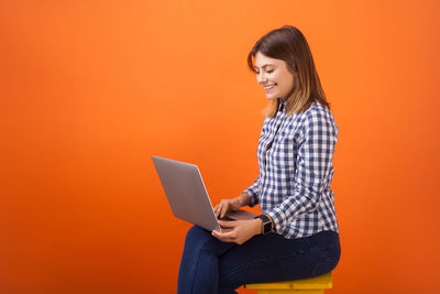 Side view of young woman using mobile phone against orange background