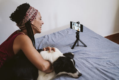 Smiling woman talking with friends on video conference over mobile phone while lying with dog on bed at home