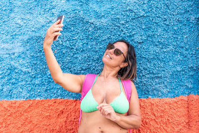 Pretty middle aged woman wearing swimsuit taking a selfie with colored wall as a background
