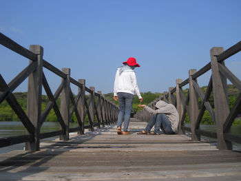 Rear view of woman giving money to beggar sitting on footbridge against clear sky