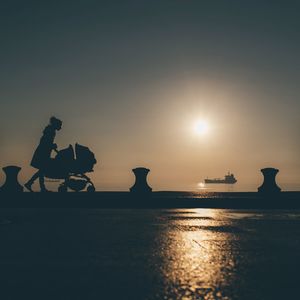 Silhouette woman pushing baby stroller on promenade during sunset