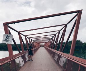 Rear view of man standing on bridge over river at lorong halus wetland against cloudy sky