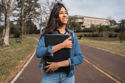 Smiling young hispanic female university student in denim clothes holding laptop and looking away while dreaming about future as spending day in green park in campus