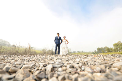 Full length of bride and bridegroom standing on stones against sky