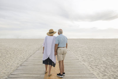 Back view of senior couple standing hand in hand on wooden boardwalk looking at horizon, liepaja, latvia