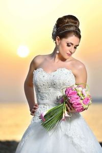 Beautiful bride holding bunch of flowers while standing at beach against sky during sunset