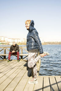 Senior woman stretching leg while man sitting at bench on pier against clear sky