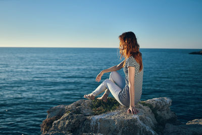 Woman sitting on rock looking at sea against clear sky