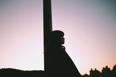 Side view of thoughtful woman leaning on pole against clear sky during sunset