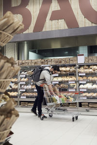 Man keeping paper bag in shopping cart while standing by bread rack at supermarket