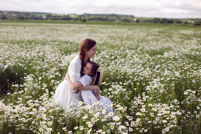 Mother with daughter in a white dress and hat stand in a daisy field in summer