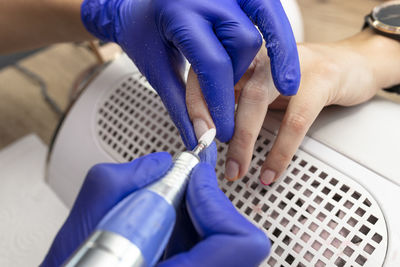 The manicurist grinds the nail plate with a milling machine in blue latex gloves.