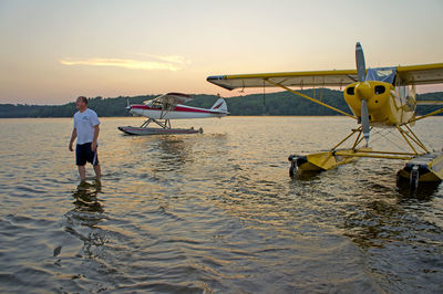 Man standing by seaplanes in water against sky during sunset