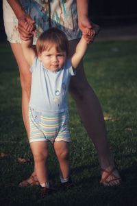 Baby boy with blonde hair and blue eyes on grass making first steps helped by parent arms