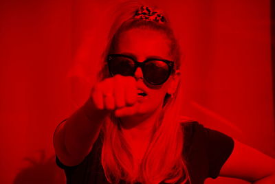 Portrait of young woman wearing sunglasses while gesturing fist against red background