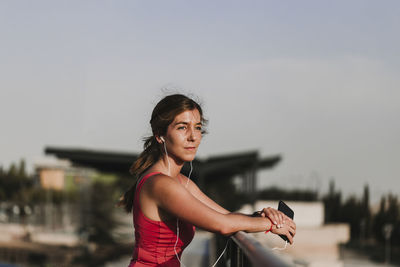 Female athlete listening music while standing by railing against sky