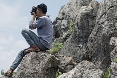 Side view of man sitting on rock while photographing from camera