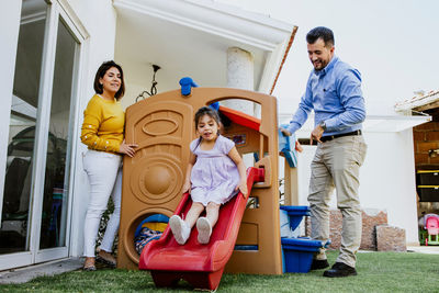 Full body of joyful little ethnic girl playing on slide while spending time in backyard with young parents on sunny day