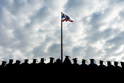 Low angle view of silhouette graduated students standing by flag against cloudy sky