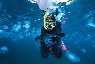 A woman in a black wetsuit is in the water with jellyfish. the jellyfish are floating around her