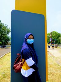 A girl wearing a blue hijab, a mask and a muslim casual tshirt standing on an empty blue billboard