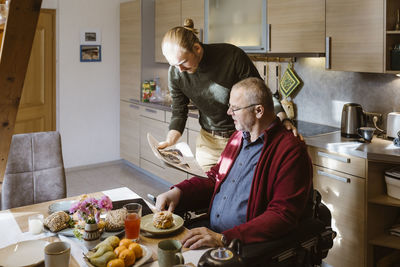 Young caretaker sharing newspaper with senior man while eating breakfast at home