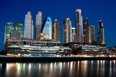 Illuminated modern buildings by river against sky in city