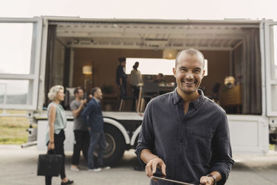 Portrait of smiling businessman holding digital tablet with colleagues and portable office truck on road in background