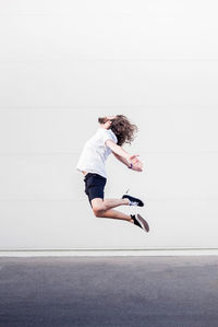 Full length of young man with arms outstretched jumping on road against wall