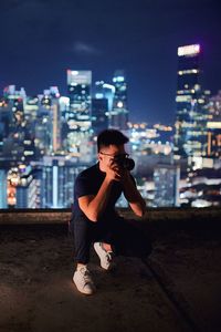 Young man sitting against illuminated buildings in city at night