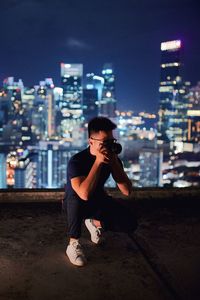 Full length of man photographing on building terrace against illuminated cityscape at night