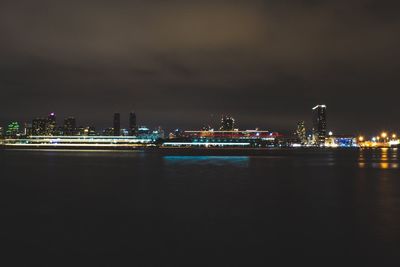 Illuminated city at waterfront against cloudy sky