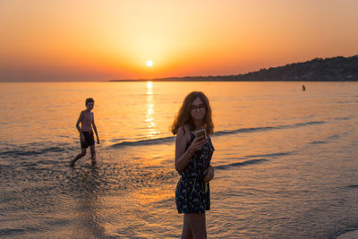 Teenage girl using phone near boy walking at beach during sunset