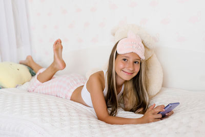 Beautiful teenage girl in pajamas is lying on a cozy bed on her stomach and surfing on the phone