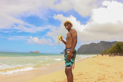 Shirtless man eating banana while standing at beach against sky