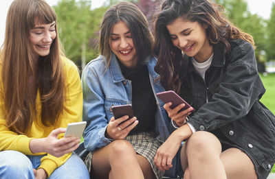 Smiling women using mobile phone while sitting together at park