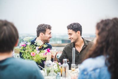 Happy man toasting drink with male friend while sitting on rooftop against sky