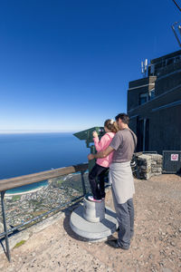 Rear view of father and daughter standing by binoculars against sea and clear blue sky