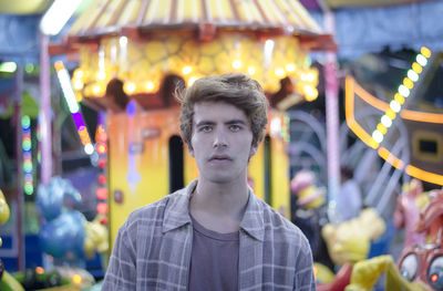 Portrait of young man standing against carousel at amusement park