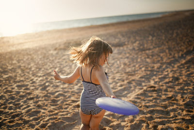 Cute european child girl in a swimsuit runs on the sand and plays frisbee on the beach. 