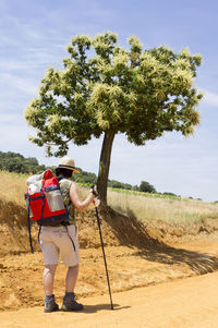 Side view of hiker in hat with backpack walking on dirt road against sky during sunny day