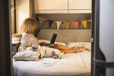 Woman using laptop while sitting with dog on bed