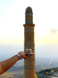 Cropped hand of woman holding cup against tower during sunset