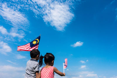 Rear view of friends holding malaysian flags while standing against blue sky