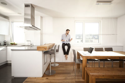 Man with coffee cup sitting on sideboard in his open plan kitchen looking at smartphone