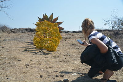 Side view of woman using mobile phone while crouching on field against clear sky during sunny day