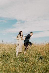 Young couple holding hands while walking on grassy field against cloudy sky