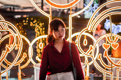 Woman looking away while standing against illuminated lighting equipment at night